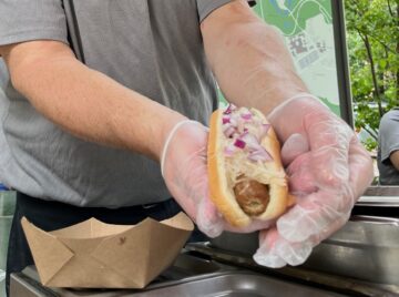 A man extends a hot dog with toppings toward the camera